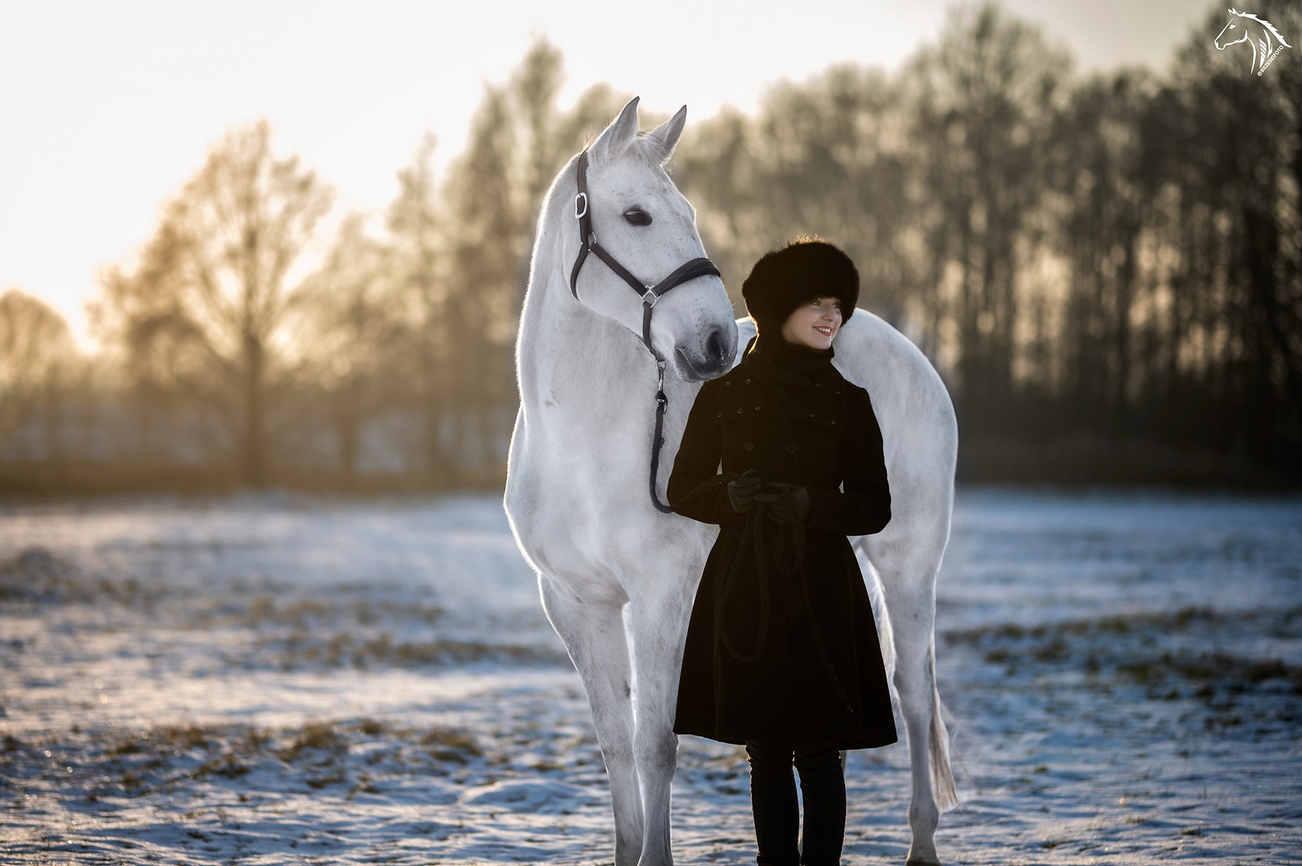 Uśmiechnięta kobieta w czarnym płaszczu i futrzanej czapce stoi obok białego konia na ośnieżonym polu, w tle drzewa oświetlone zachodzącym słońcem.