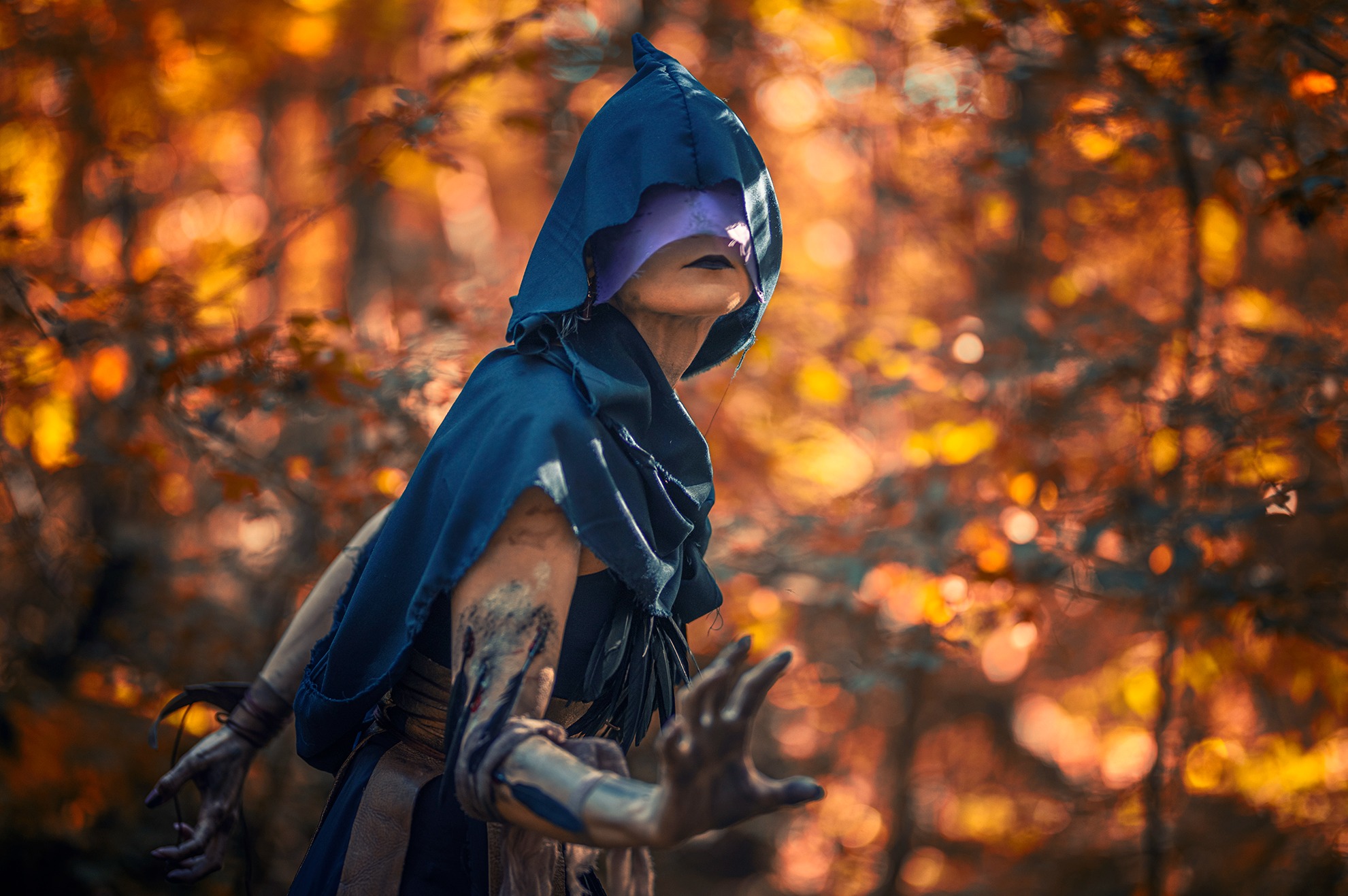 Kobieta w niebieskim kapturze i masce na tle jesiennego lasu, z rękami wyciągniętymi w kierunku widza, makijaż w stylu fantasy.