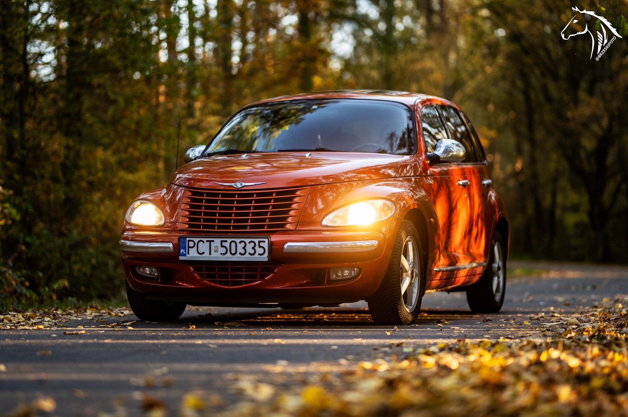 Pomarańczowy Chrysler PT Cruiser na asfalcie pokrytym jesiennymi liśćmi, światła włączone, tło z drzewami w jesiennych barwach.