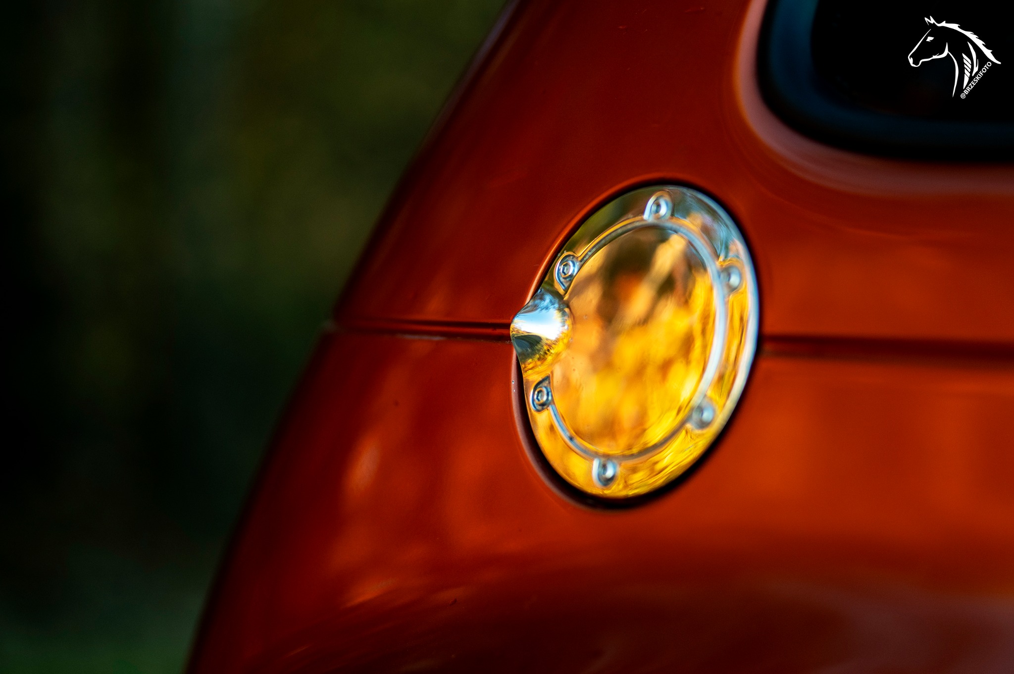 Chromowana klapka wlewu paliwa na karoserii samochodu w kolorze pomarańczowym, z widocznym odbiciem otoczenia i logotypem fotografa w prawym górnym rogu.