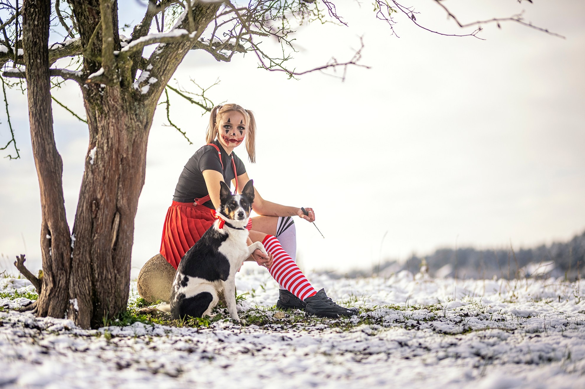 Młoda kobieta z makijażem klauna, w czerwonej spódnicy i podkolanówkach w paski, siedzi obok drzewa na śniegu z czarno-białym psem w muszce.