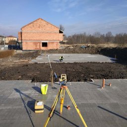 GRUPA K7 - Praca na budowie: żółty teodolit na trójnogu ustawiony na świeżym fundamencie, w tle budynek w stanie surowym otwartym z czerwonej cegły i pracujący człowiek w kamizelce odblaskowej.