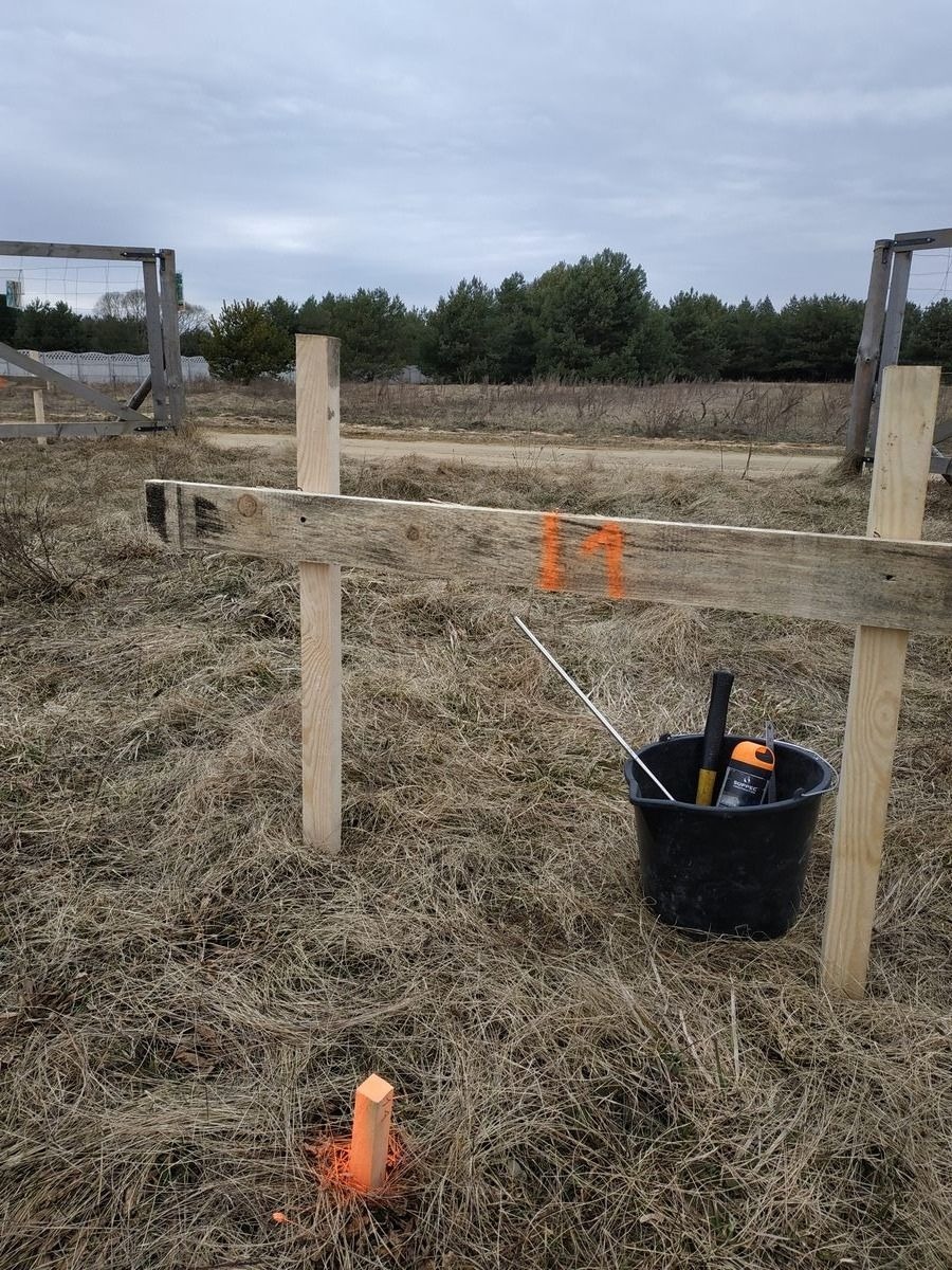 Wytyczanie granic działki: drewniane słupki z pomarańczowymi oznaczeniami w trawie, czarne wiadro z narzędziami w tle, pochmurne niebo.