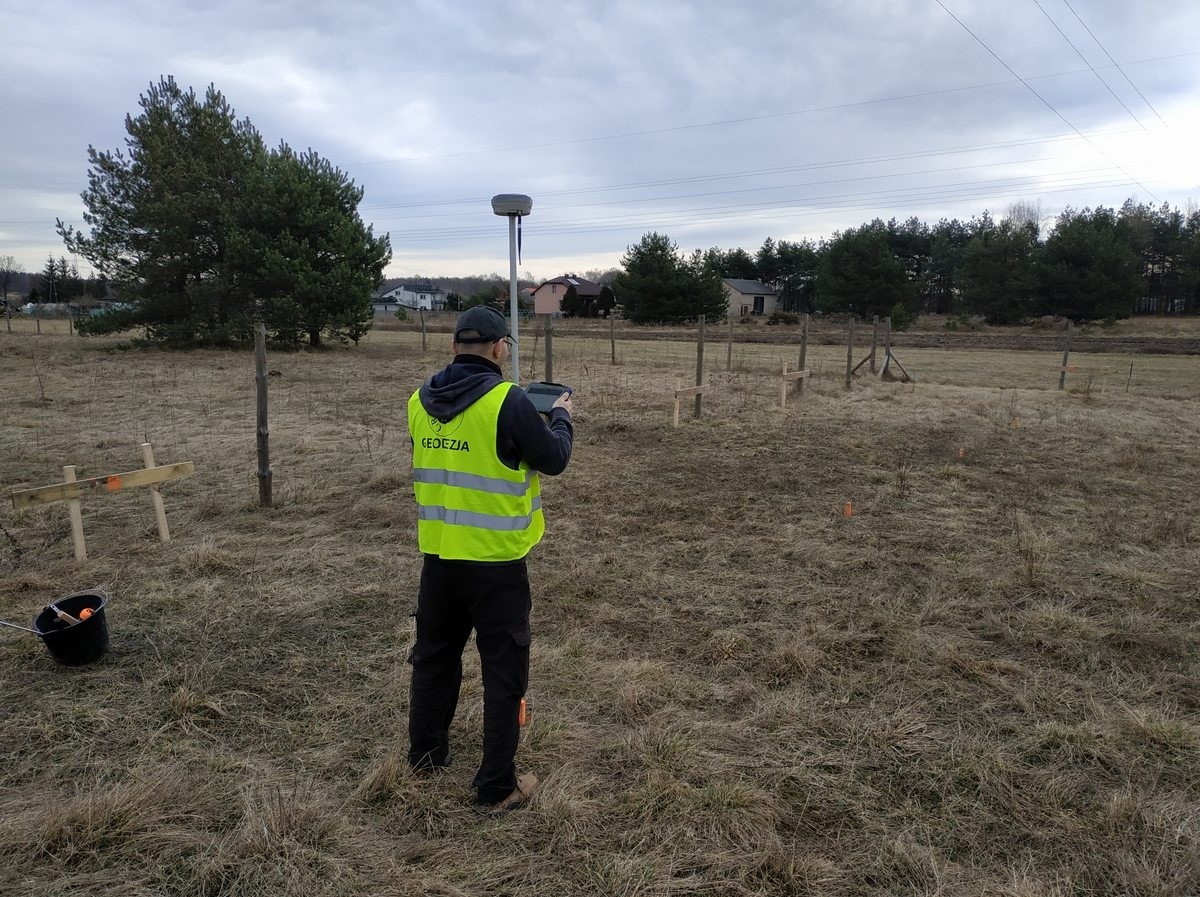 Mężczyzna w kamizelce odblaskowej z napisem 'GEODEZJA' obsługuje urządzenie pomiarowe GPS na tle pola z wbitymi palikami i drzewami.