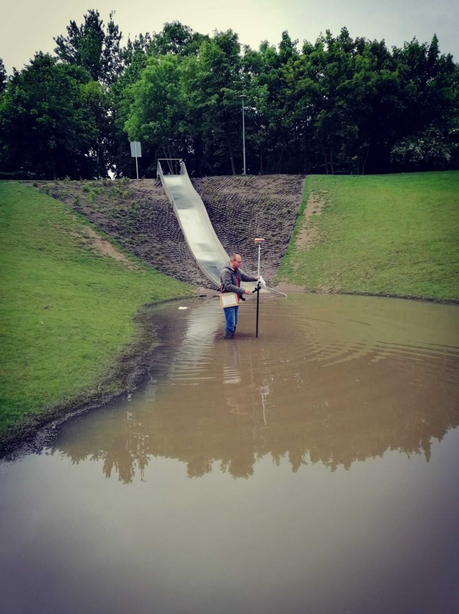 Człowiek w gumowych butach stoi w wodzie, trzymając tyczkę geodezyjną z odbiornikiem GPS, w tle zjeżdżalnia na wzgórzu i drzewa.