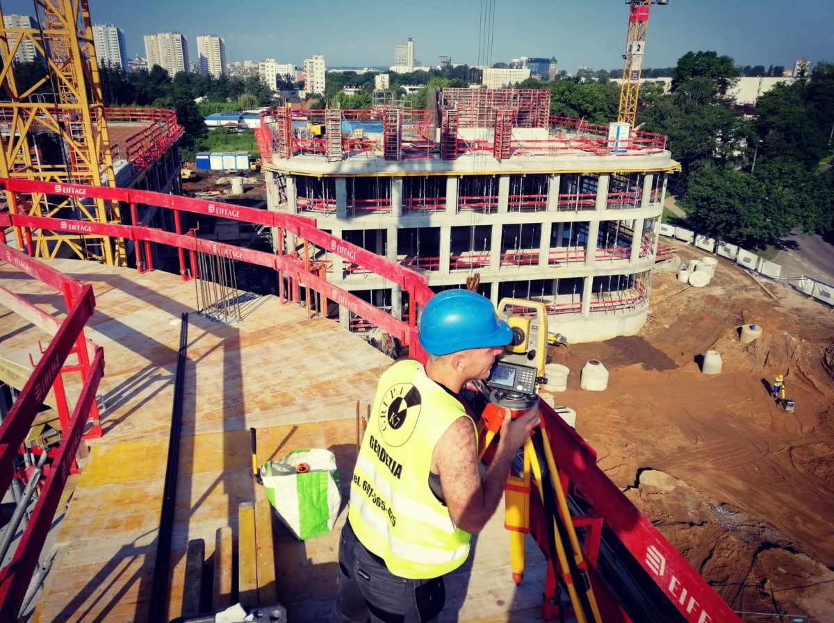 Geodeta w niebieskim kasku i żółtej kamizelce, z logo firmy, mierzy poziomicą na budowie wielopiętrowego budynku w słoneczny dzień, z widokiem na panoramę miasta w tle.