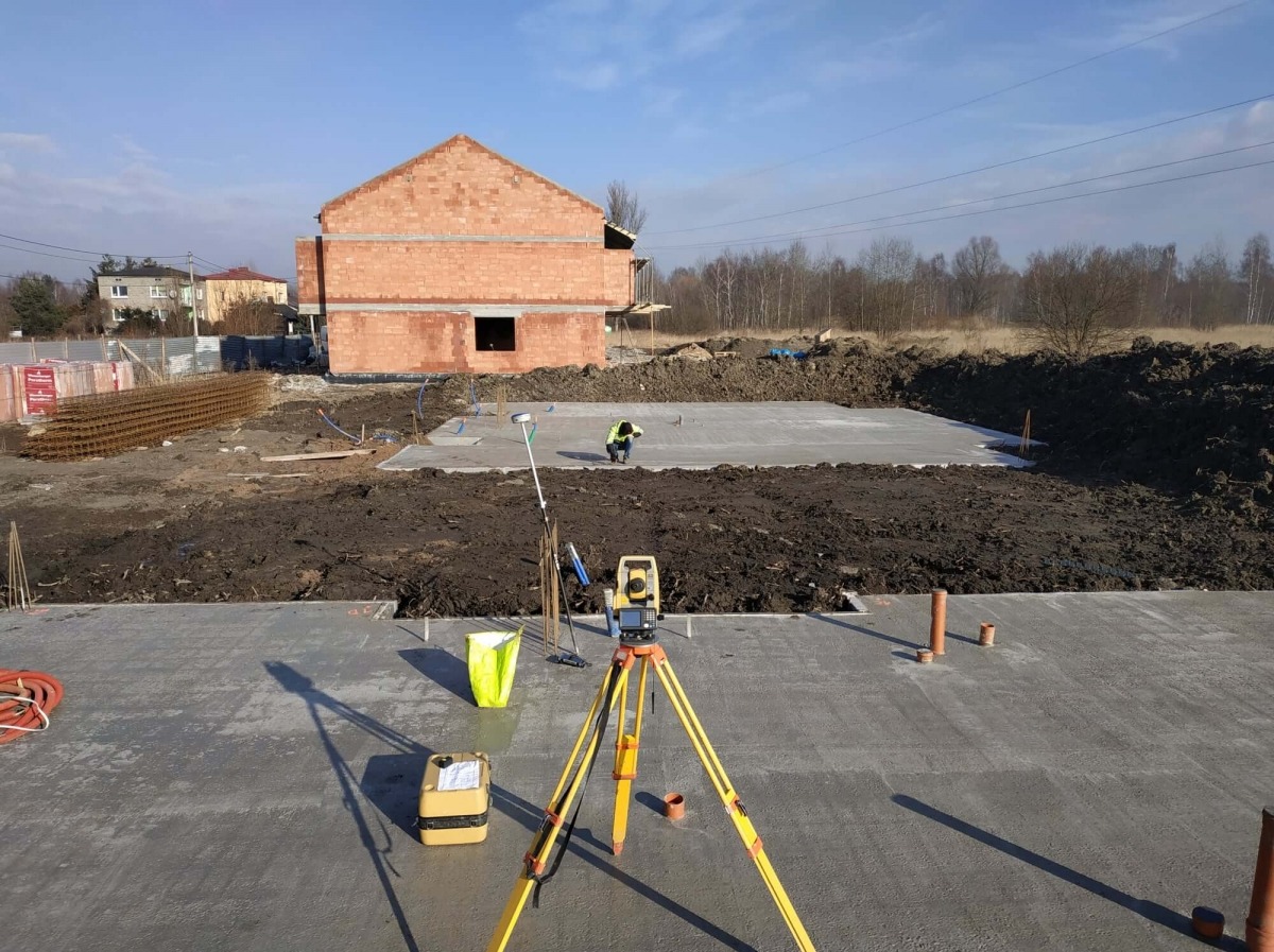 Praca na budowie: żółty teodolit na trójnogu ustawiony na świeżym fundamencie, w tle budynek w stanie surowym otwartym z czerwonej cegły i pracujący człowiek w kamizelce odblaskowej.