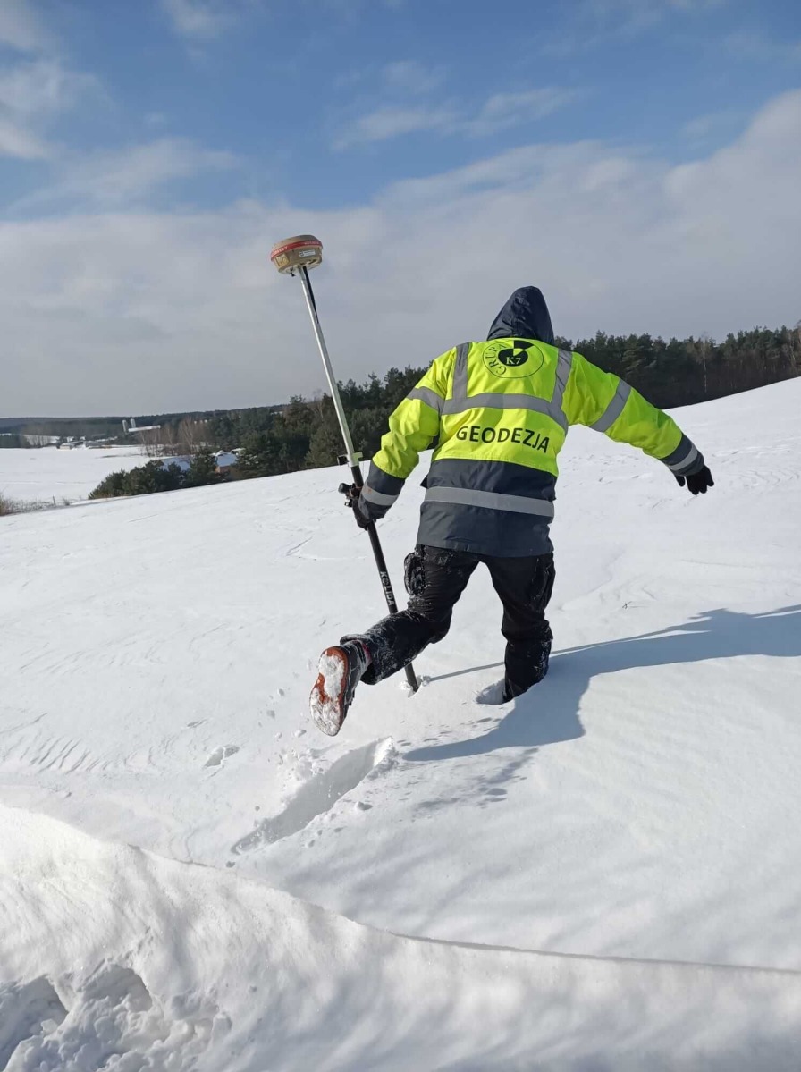Osoba w odblaskowej kurtce z napisem GEODEZJA, niosąca tyczkę geodezyjną z odbiornikiem GPS, brodzi w głębokim śniegu na wzgórzu, w tle las i błękitne niebo.