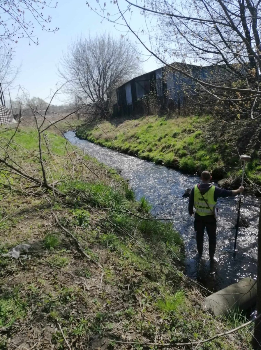 Mężczyzna w żółtej kamizelce brodzi w płytkim strumieniu, trzymając tyczkę geodezyjną; w tle zarośnięty brzeg i blaszany budynek.