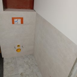 Mateusz Krzemiński - Wnęka w łazience z zamontowaną instalacją podtynkową WC, widoczne płytki ścienne w odcieniach beżu, żółte elementy instalacji i pomarańczowa obudowa stelaża JOMO.