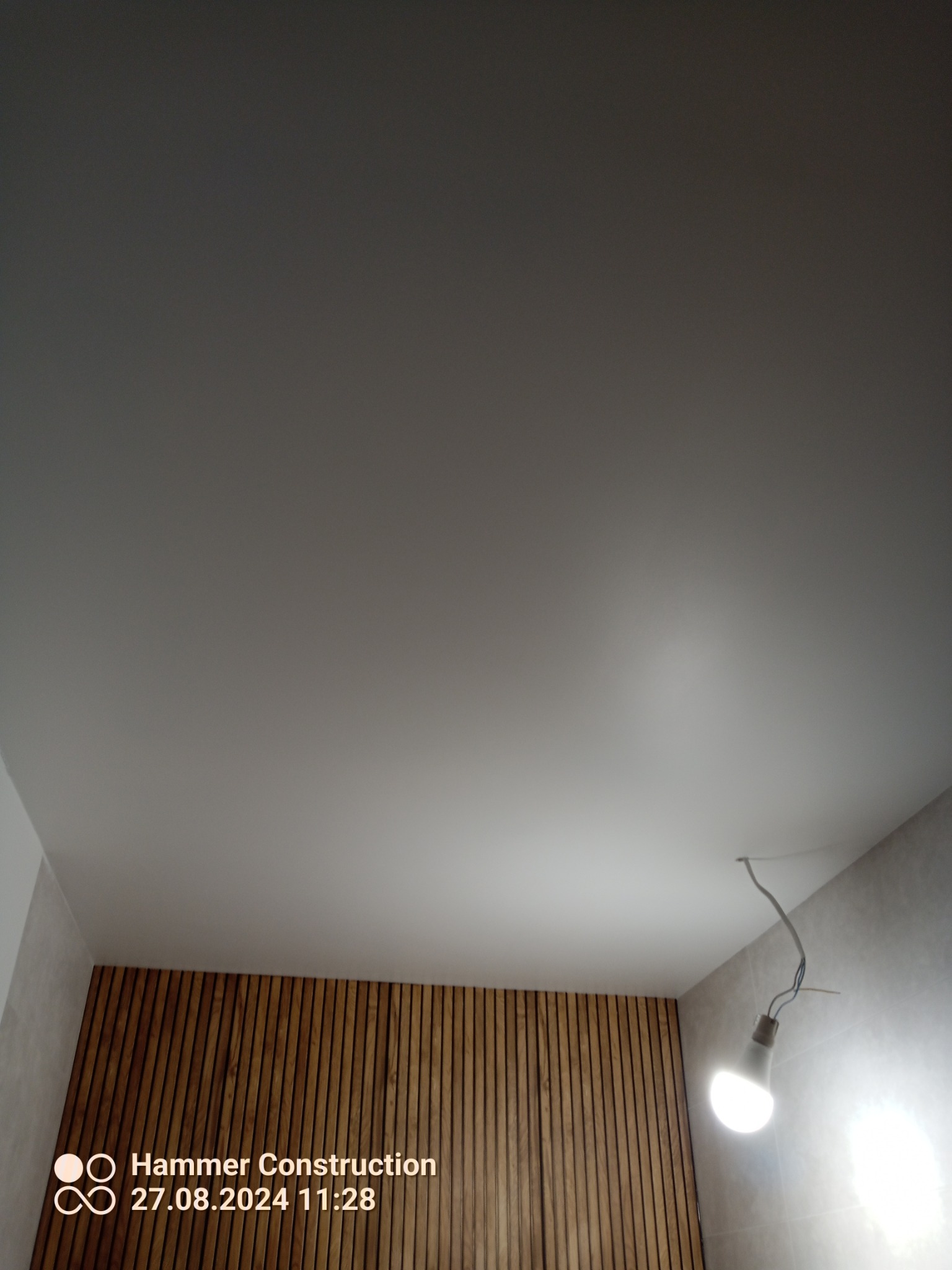 Gładki, biały sufit z zamontowaną lampą wiszącą na kablu, obok ściana z drewnianymi panelami i fragment ściany wykończonej płytkami.