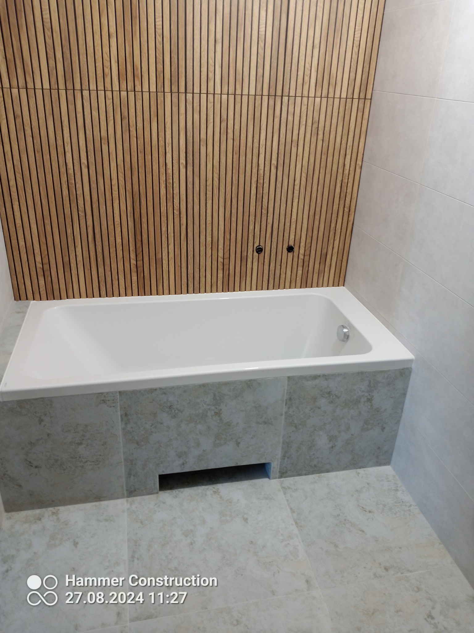 Nowoczesna wanna obudowana płytkami w odcieniach beżu, z drewnianą ścianą z pionowych paneli i jasnymi płytkami na ścianach, tworząca minimalistyczną aranżację łazienki.