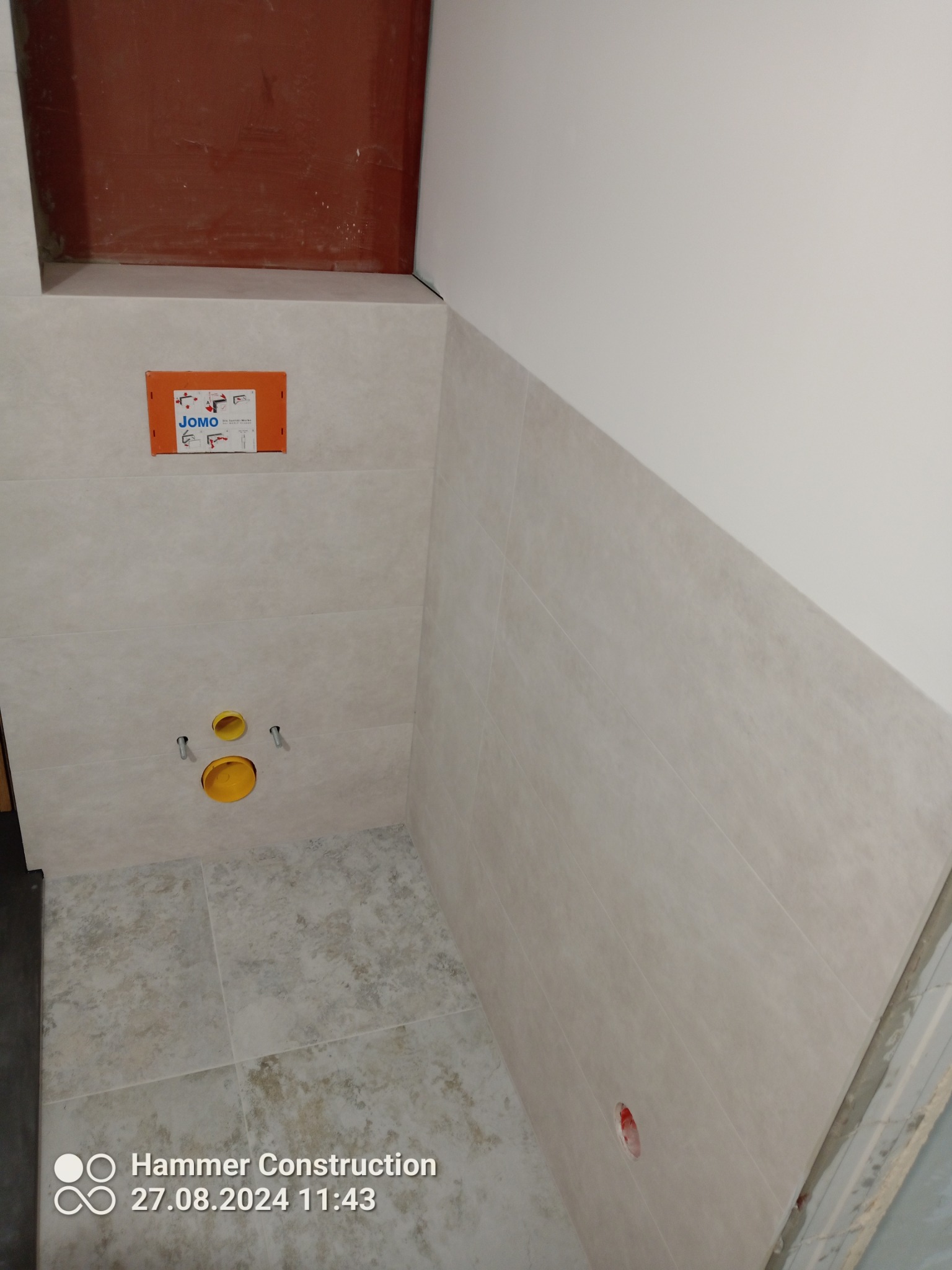 Wnęka w łazience z zamontowaną instalacją podtynkową WC, widoczne płytki ścienne w odcieniach beżu, żółte elementy instalacji i pomarańczowa obudowa stelaża JOMO.