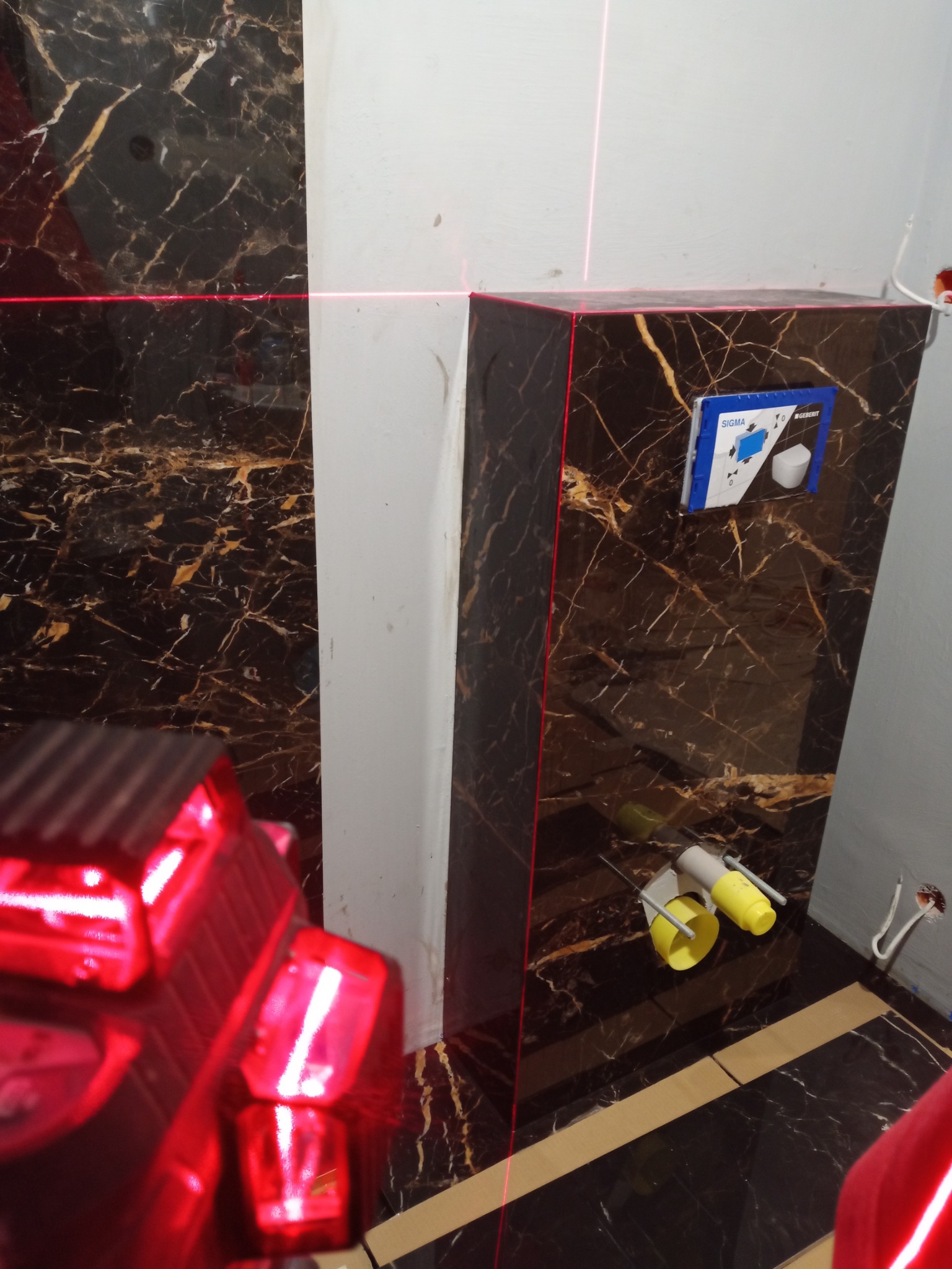 Montaż podtynkowego stelaża WC na tle marmurowej okładziny, z użyciem lasera krzyżowego do precyzyjnego ustawienia.