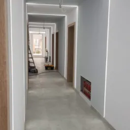 Długi korytarz w trakcie prac wykończeniowych, z zamontowanym oświetleniem LED wzdłuż sufitu i ścian, widoczne narzędzia i drabina w tle, szare płytki na podłodze, drewniane ościeżnice drzwiowe.