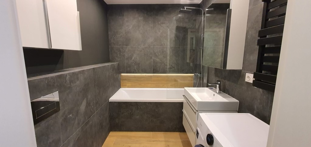 Nowoczesna łazienka w Kczewie z szarymi płytkami imitującymi beton, białą wanną z drewnianym panelem, umywalką nablatową i pralką, po remoncie.