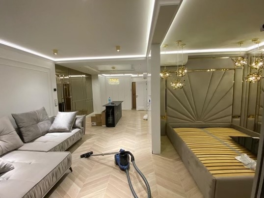 Wnętrze luksusowego apartamentu po remoncie z parkietem w jodełkę, szarą sofą, wyspą kuchenną, łóżkiem z tapicerowanym zagłówkiem w stylu art deco i odkurzaczem na podłodze.