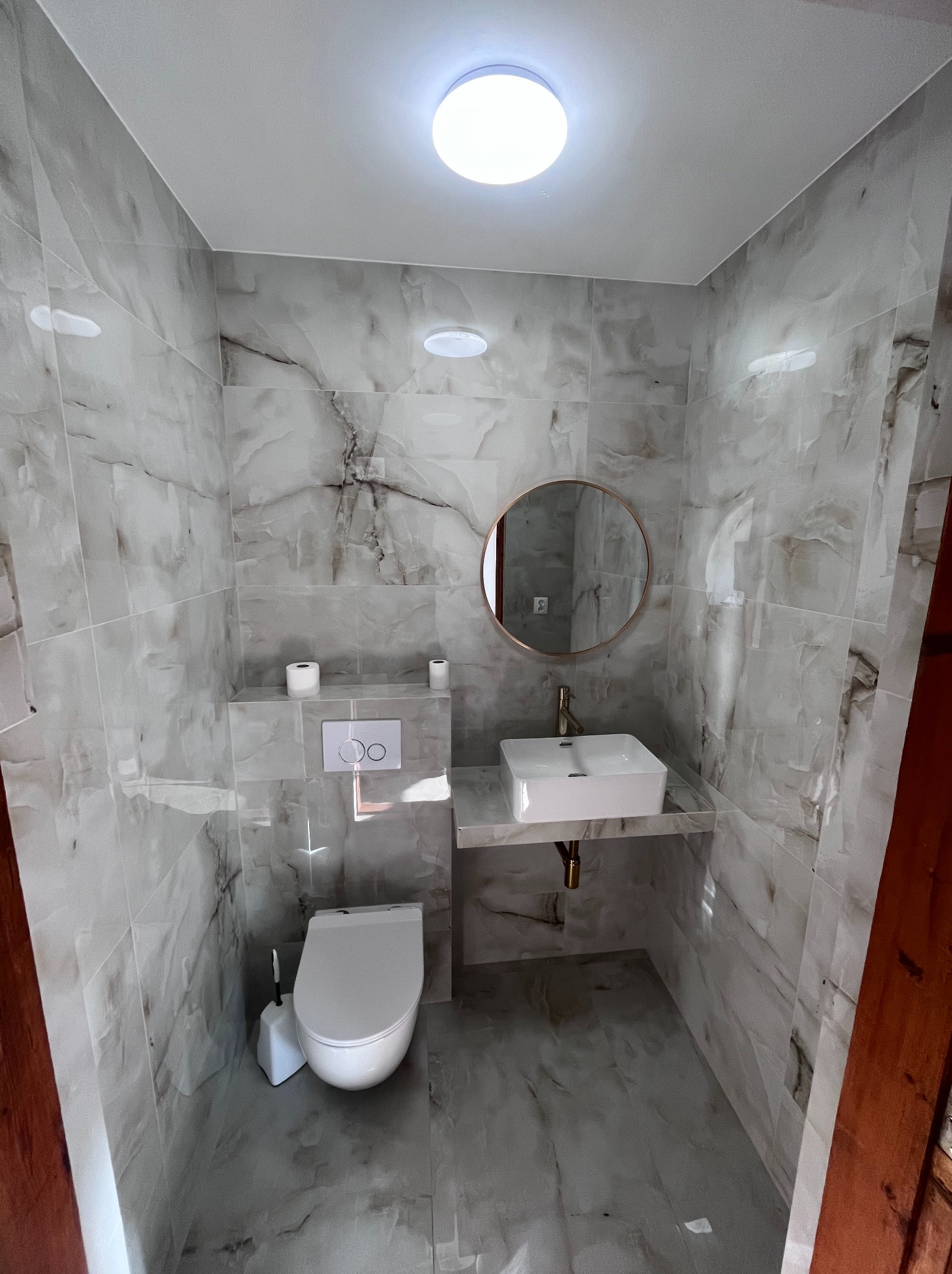 Wykończona łazienka w szaro-białych marmurowych płytkach, z wiszącą toaletą, umywalką na blacie, okrągłym lustrem w złotej ramie i oświetleniem sufitowym.