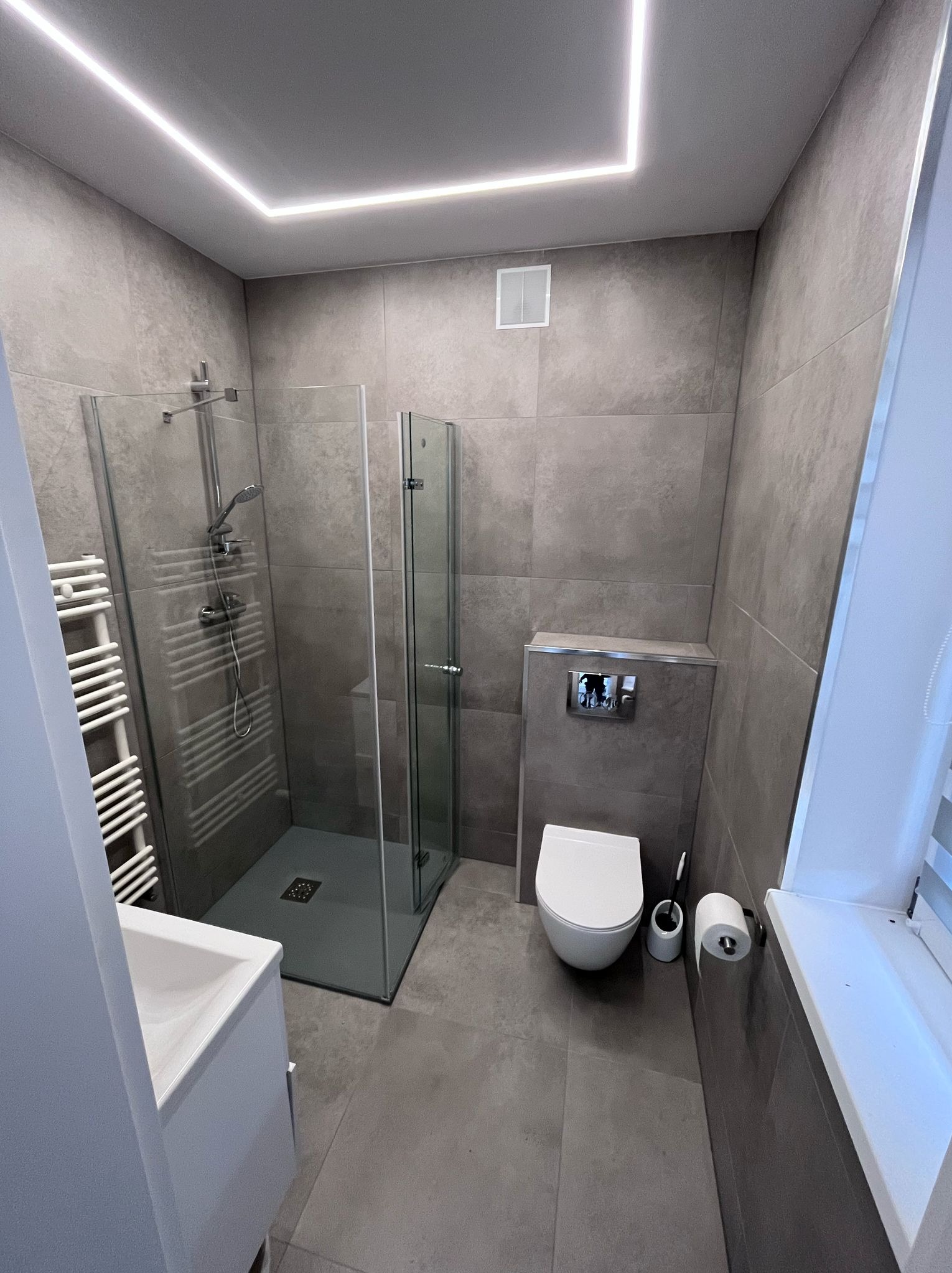 Nowoczesna łazienka z szarymi płytkami, kabiną prysznicową ze szklanymi drzwiami, podwieszaną toaletą i oświetleniem LED w suficie.