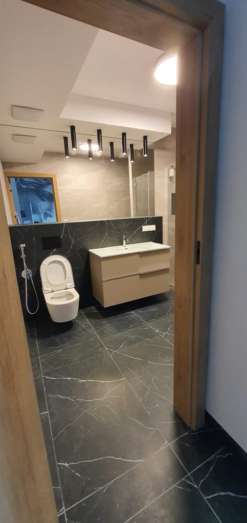 Nowoczesna łazienka z czarną marmurową podłogą i ścianą za toaletą i umywalką, beżowa szafka pod umywalką, wisząca toaleta, czarne oświetlenie punktowe na suficie, widok zza drewnianej ościeżnicy.