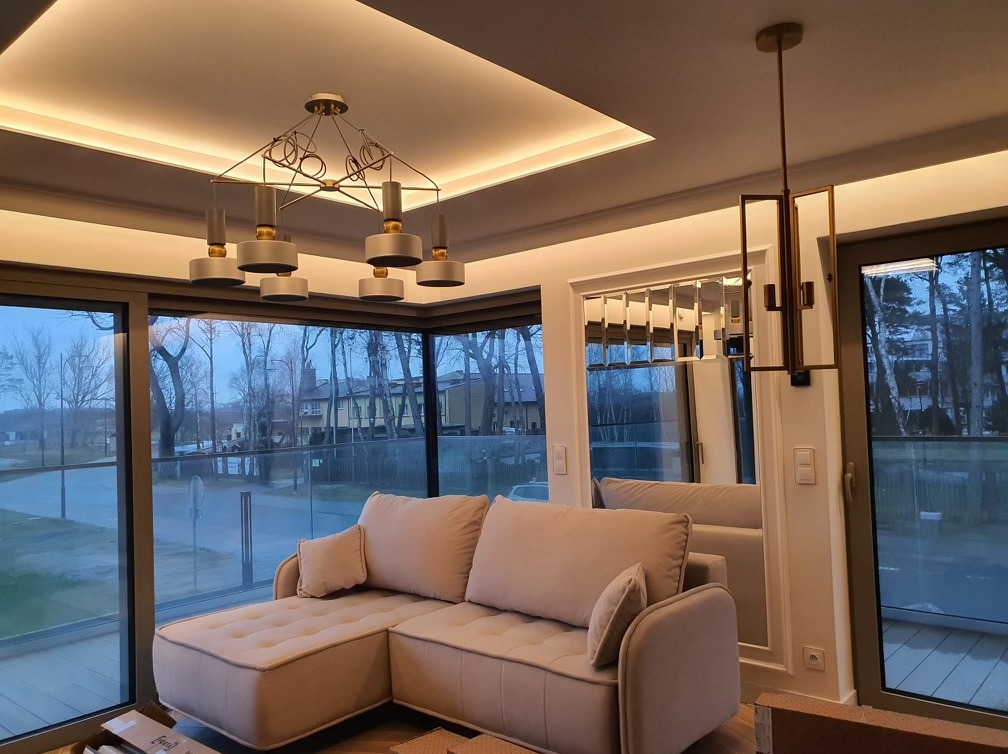 Nowoczesny salon z narożnikiem w odcieniach beżu, oświetleniem LED na suficie podwieszanym, designerskimi lampami i dużymi oknami z widokiem na zewnątrz.