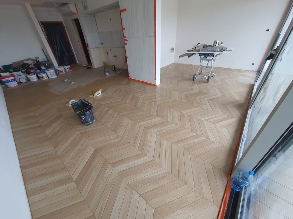 Układanie parkietu w jodełkę w jasnym pomieszczeniu, widoczny kubeł z narzędziami i wózek z materiałami, ściany przygotowane do malowania.