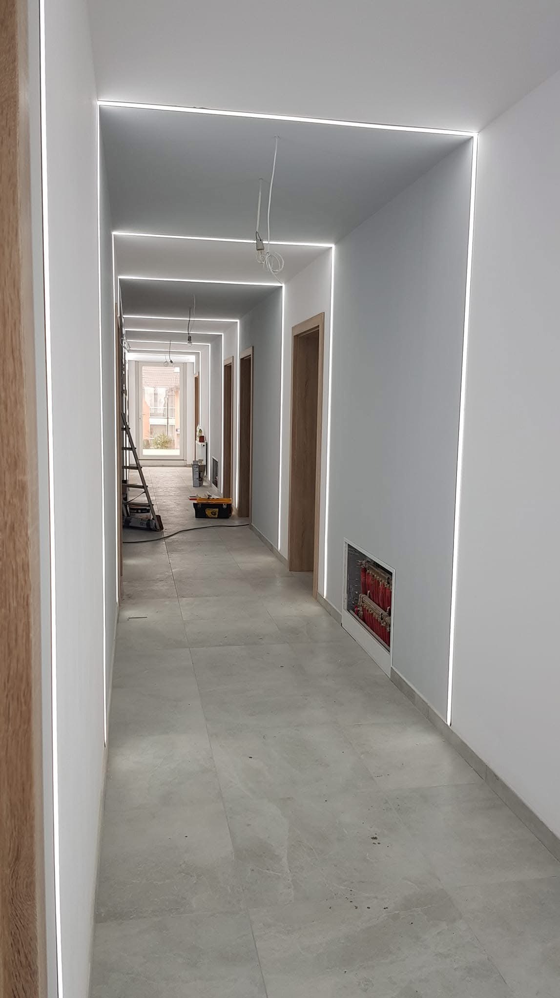 Długi korytarz w trakcie prac wykończeniowych, z zamontowanym oświetleniem LED wzdłuż sufitu i ścian, widoczne narzędzia i drabina w tle, szare płytki na podłodze, drewniane ościeżnice drzwiowe.
