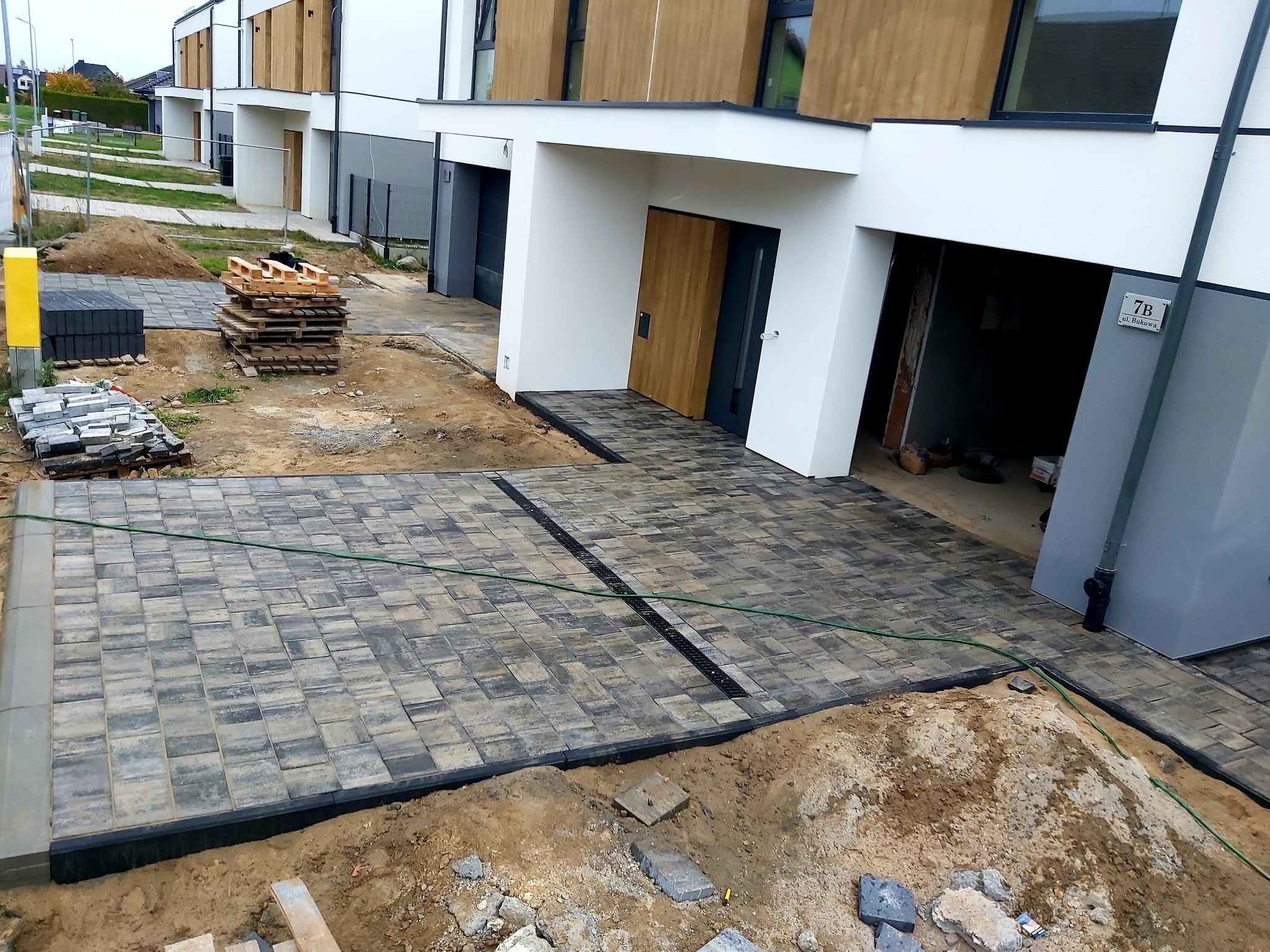 Ułożona kostka brukowa w odcieniach szarości przed szeregiem nowoczesnych domów z drewnianymi elementami, widoczne palety kostki brukowej i niezakończone prace ziemne.