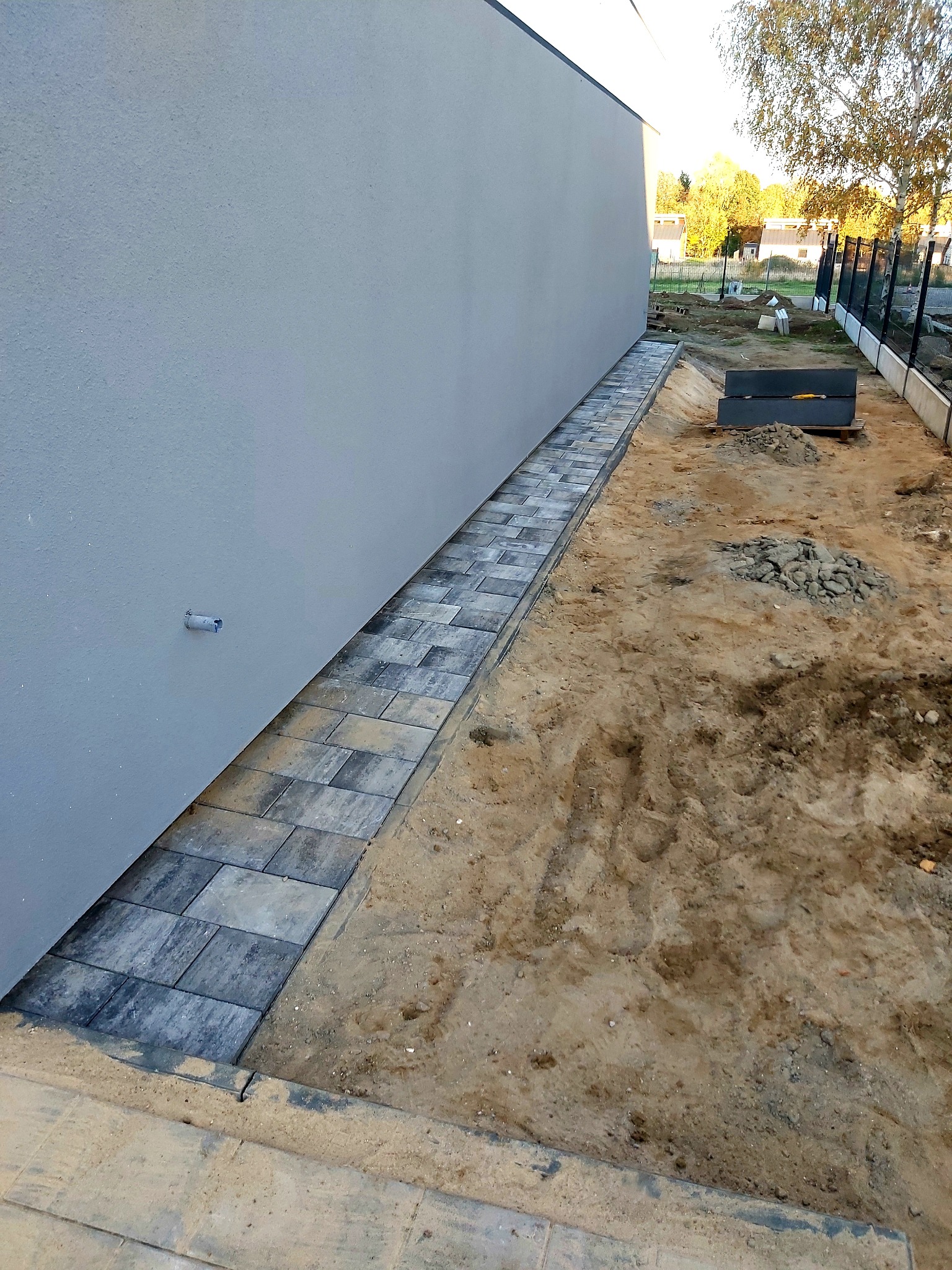 Ułożona ścieżka z kostki brukowej przylegająca do ściany budynku, widoczny teren przygotowany pod dalsze prace, w tle drzewa i ogrodzenie.
