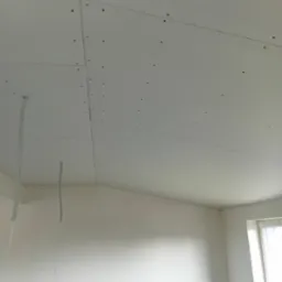 Sufit podwieszany z płyt kartonowo-gipsowych, widoczne łączenia i wkręty, wystające rury instalacyjne, surowy stan wykończenia wnętrza z oknem w tle.