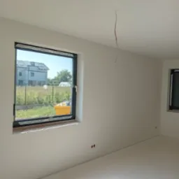 Wnętrze pokoju po świeżym malowaniu na biało, widoczne dwa okna w czarnych ramach, wystający kabel ze ściany, oraz żółty pojemnik za oknem.