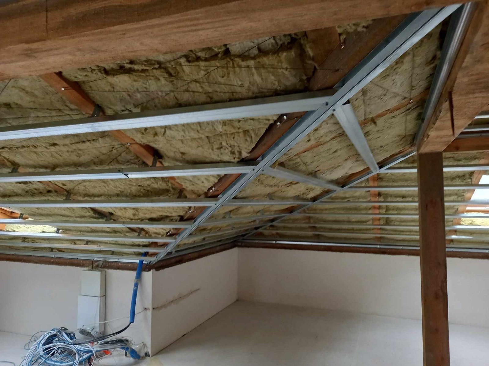 Stelaż metalowy pod płyty gipsowo-kartonowe zamontowany do drewnianej konstrukcji dachu z widoczną izolacją z wełny mineralnej, przygotowany do montażu płyt, z przewodami elektrycznymi leżącymi...
