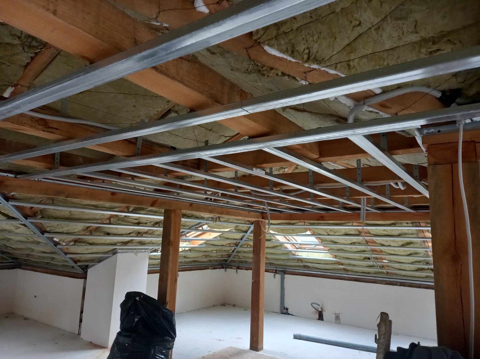 Montaż metalowej konstrukcji sufitu podwieszanego w poddaszu z widoczną izolacją termiczną, drewnianymi belkami stropowymi i fragmentem ściany po lewej stronie oraz słupami nośnymi po prawej.
