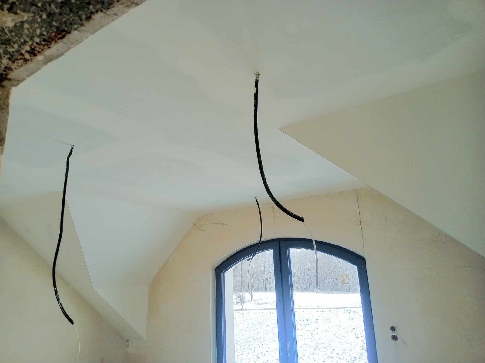 Pokój na poddaszu w trakcie wykańczania: świeżo położona gładź na skośnym suficie, wystające przewody elektryczne, okno łukowe z widokiem na zimowy krajobraz.