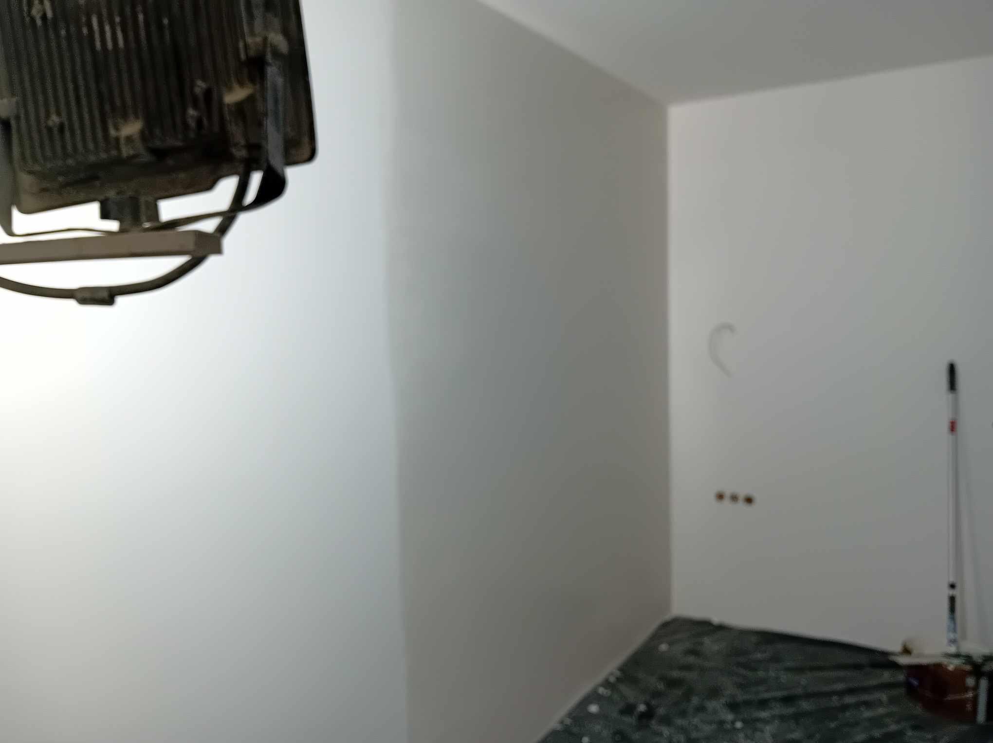 Świeżo pomalowane, białe ściany w rogu pokoju, zabezpieczone folią malarską na podłodze, z widoczną lampą halogenową i wałkiem malarskim.