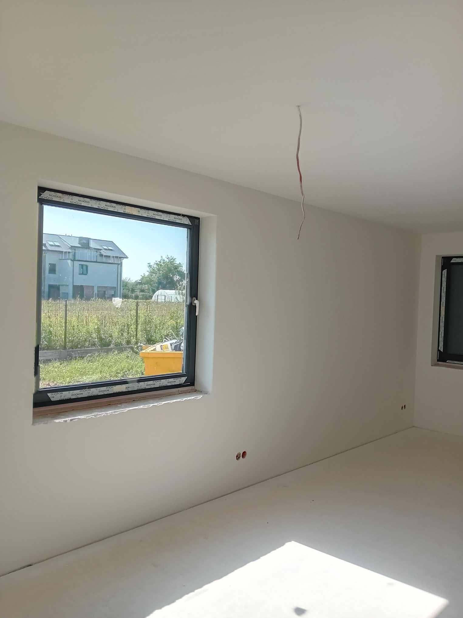 Jasne, świeżo pomalowane wnętrze pokoju z oknem z widokiem na ogród i dom w tle; widoczne wystające przewody elektryczne z sufitu i żółty pojemnik na parapecie.