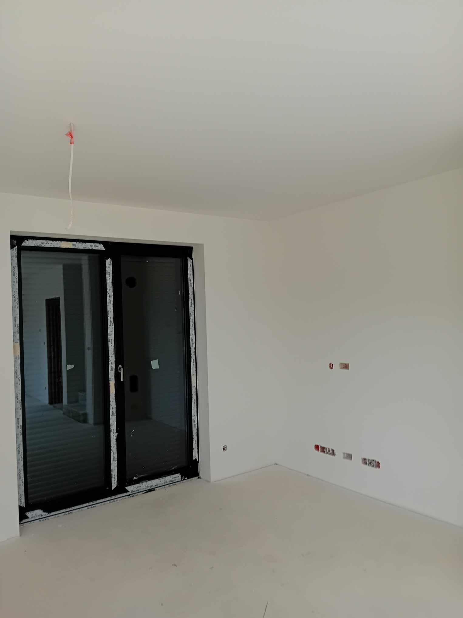 Świeżo pomalowane na biało wnętrze z dużymi przesuwnymi drzwiami balkonowymi, widoczne puste gniazdka elektryczne i wystające kable z sufitu.