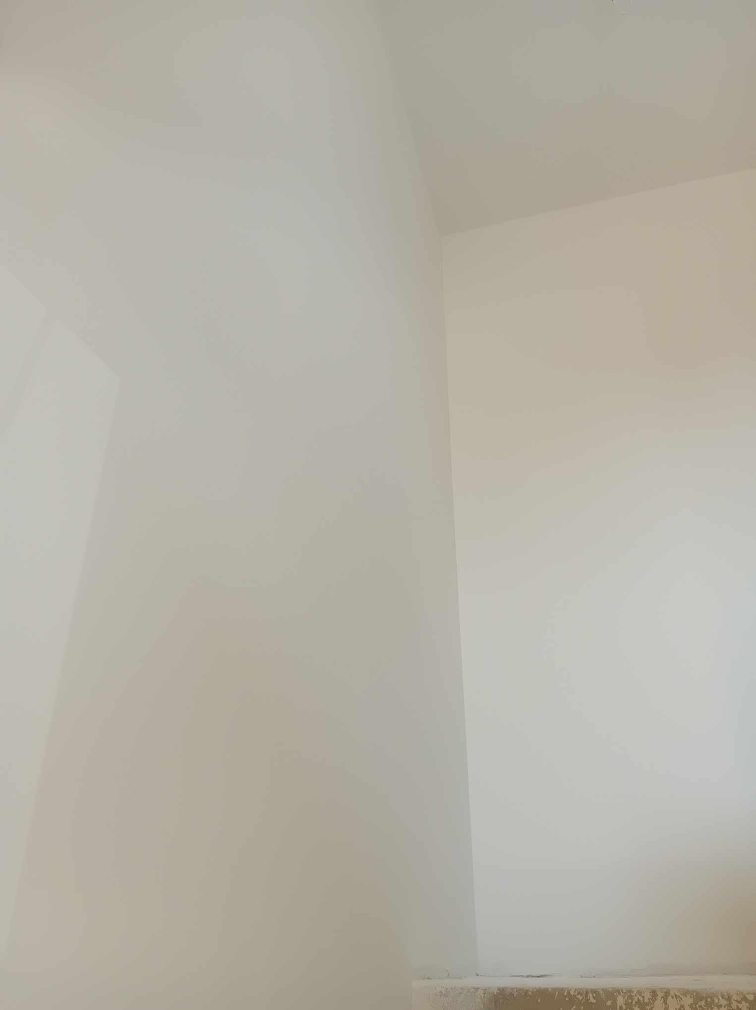 Świeżo pomalowany narożnik pomieszczenia z widocznym przejściem między ścianą a sufitem, na dole widoczne zabezpieczenia podłogi.