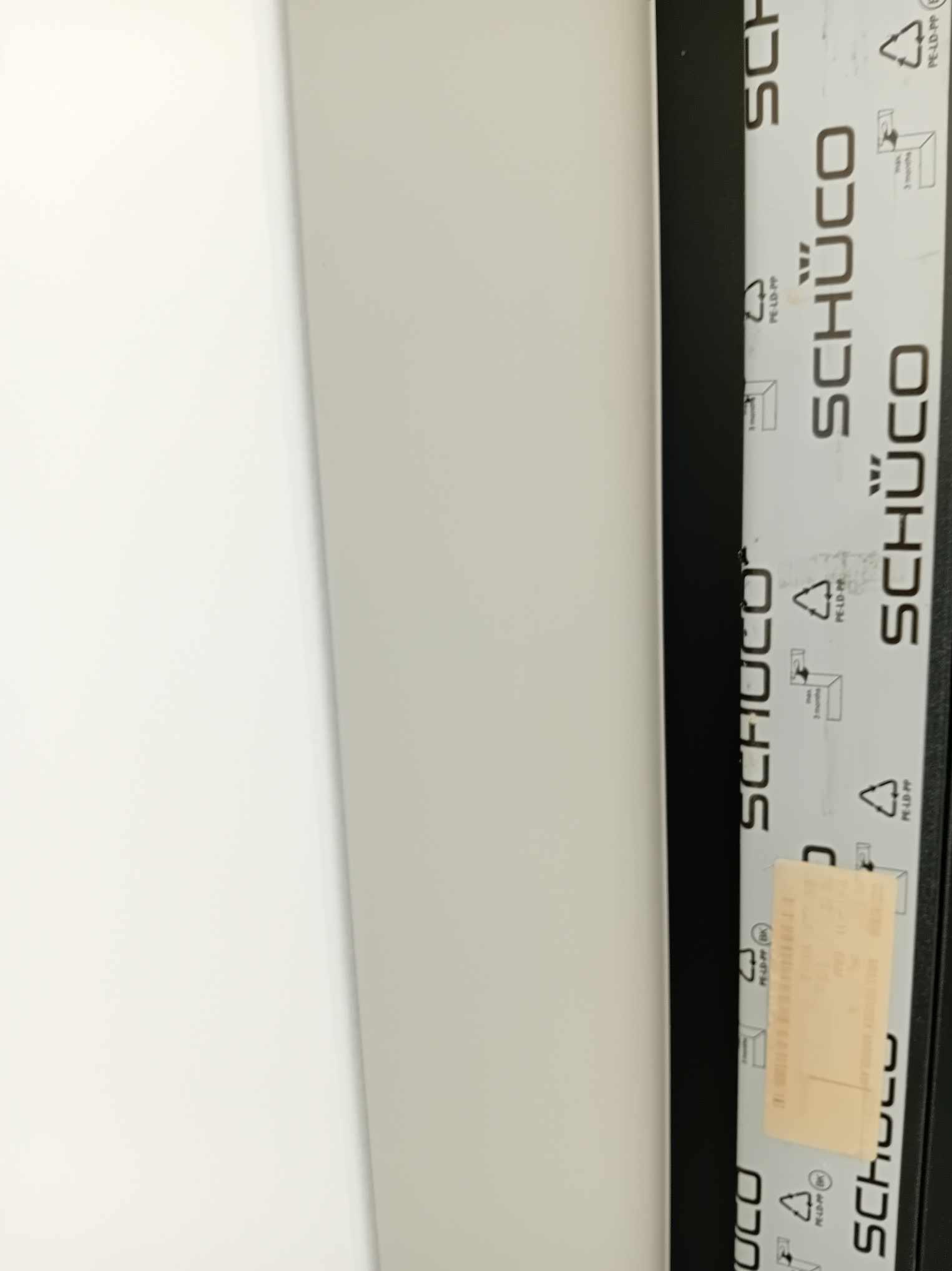 Fragment okna PCV marki Schüco z widocznymi oznaczeniami producenta i kodami recyklingu na folii ochronnej.