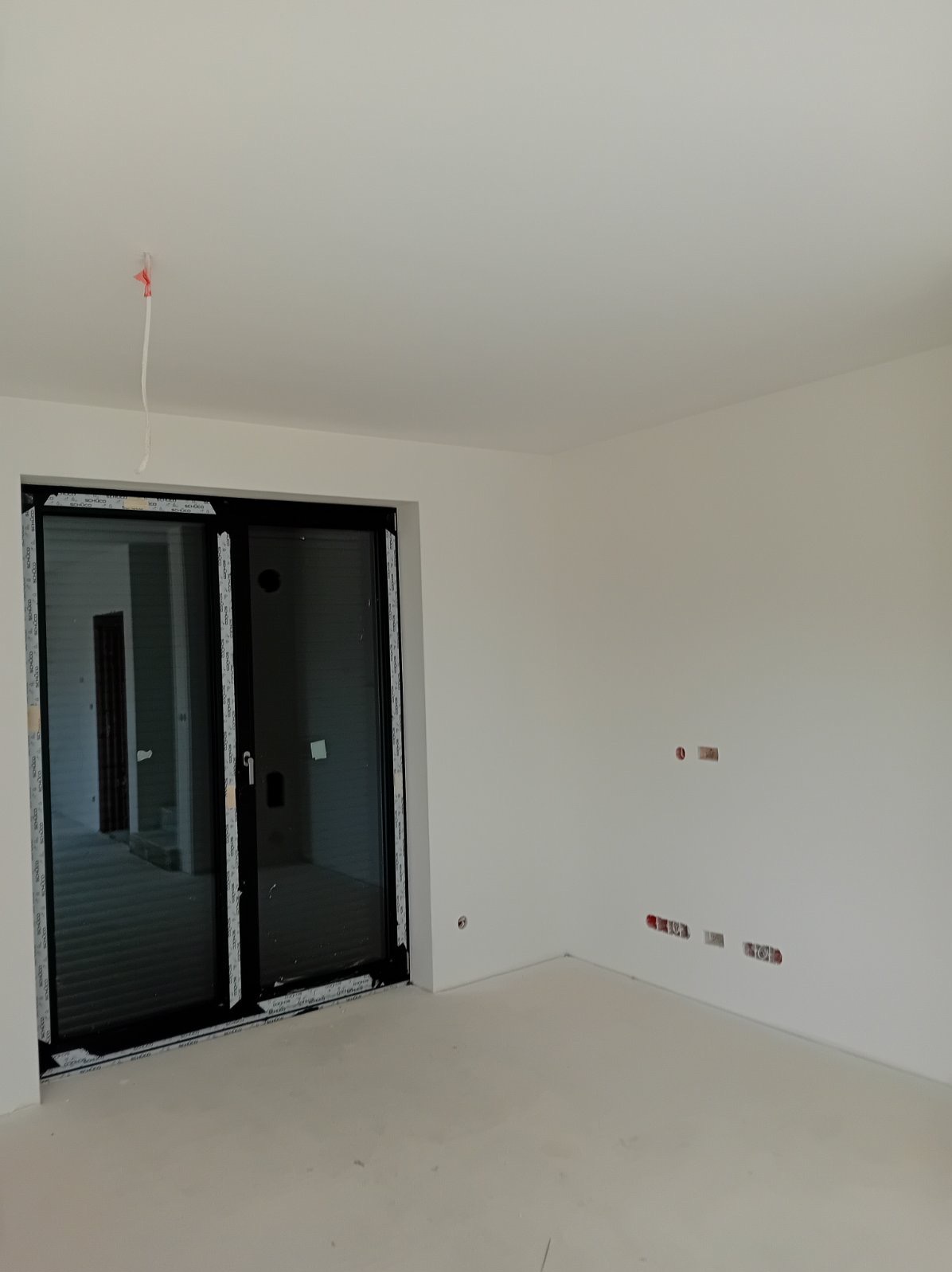 Pusty, świeżo wykończony pokój z białymi ścianami, czarnymi przesuwnymi drzwiami balkonowymi i widocznymi puszkami elektrycznymi w ścianie.