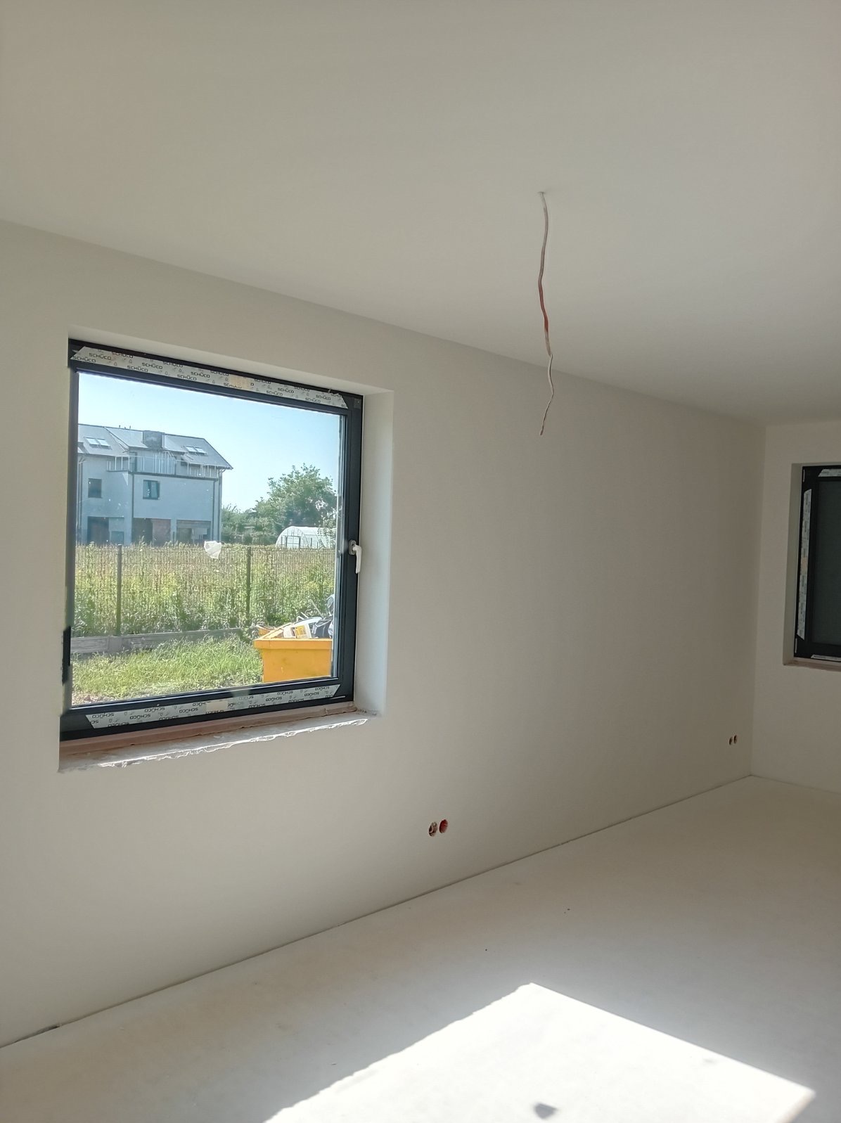 Wnętrze pokoju po świeżym malowaniu na biało, widoczne dwa okna w czarnych ramach, wystający kabel ze ściany, oraz żółty pojemnik za oknem.
