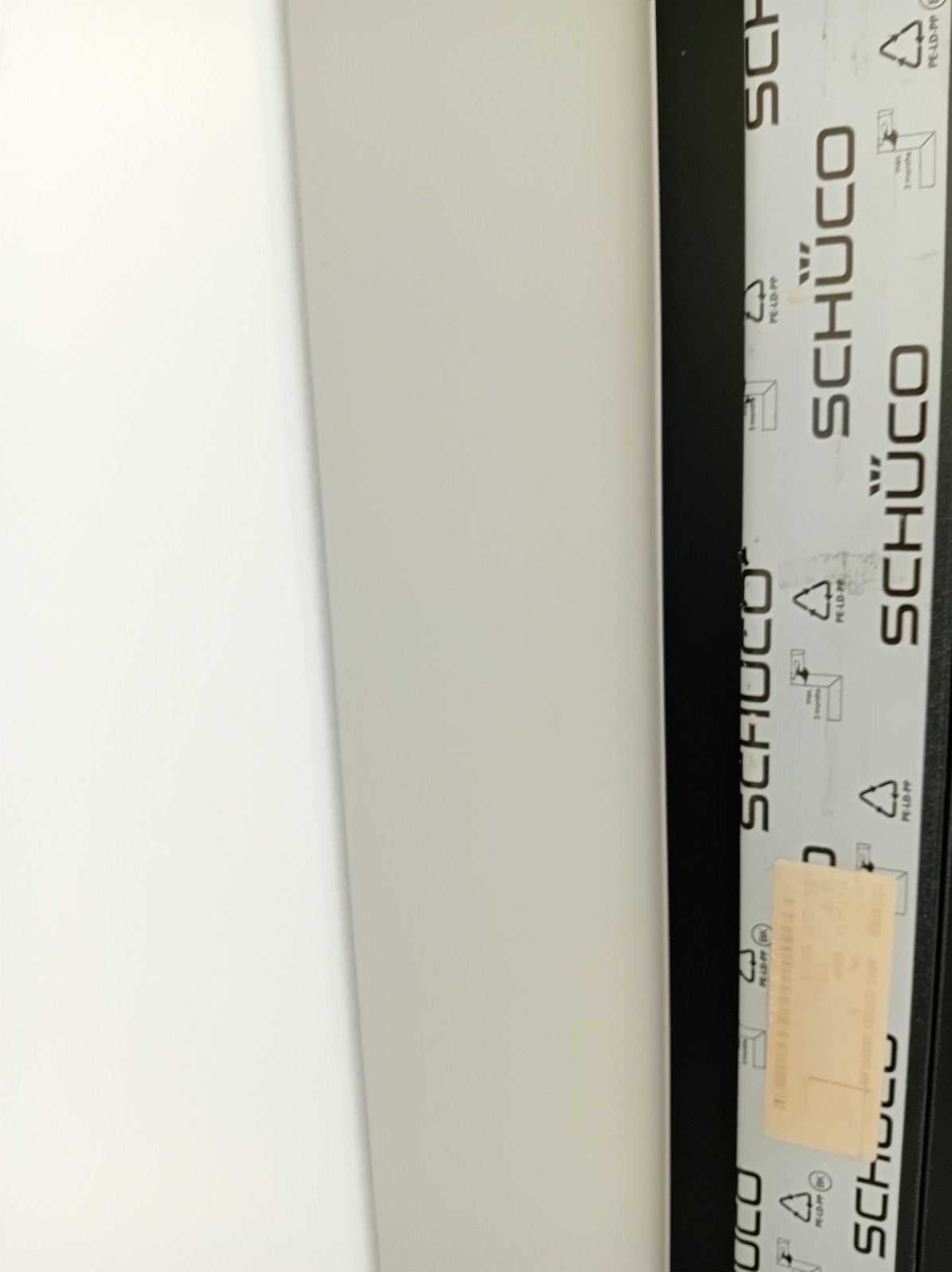 Fragment okna PCV marki Schuco, z widoczną folią ochronną i naklejką informacyjną na ramie.