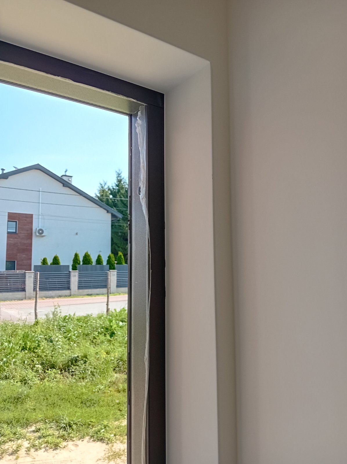 Nowo osadzone okno z brązową ramą, zabezpieczone folią ochronną, widoczne świeżo pomalowane białe ściany wewnątrz pomieszczenia i widok na dom oraz zieleń na zewnątrz.