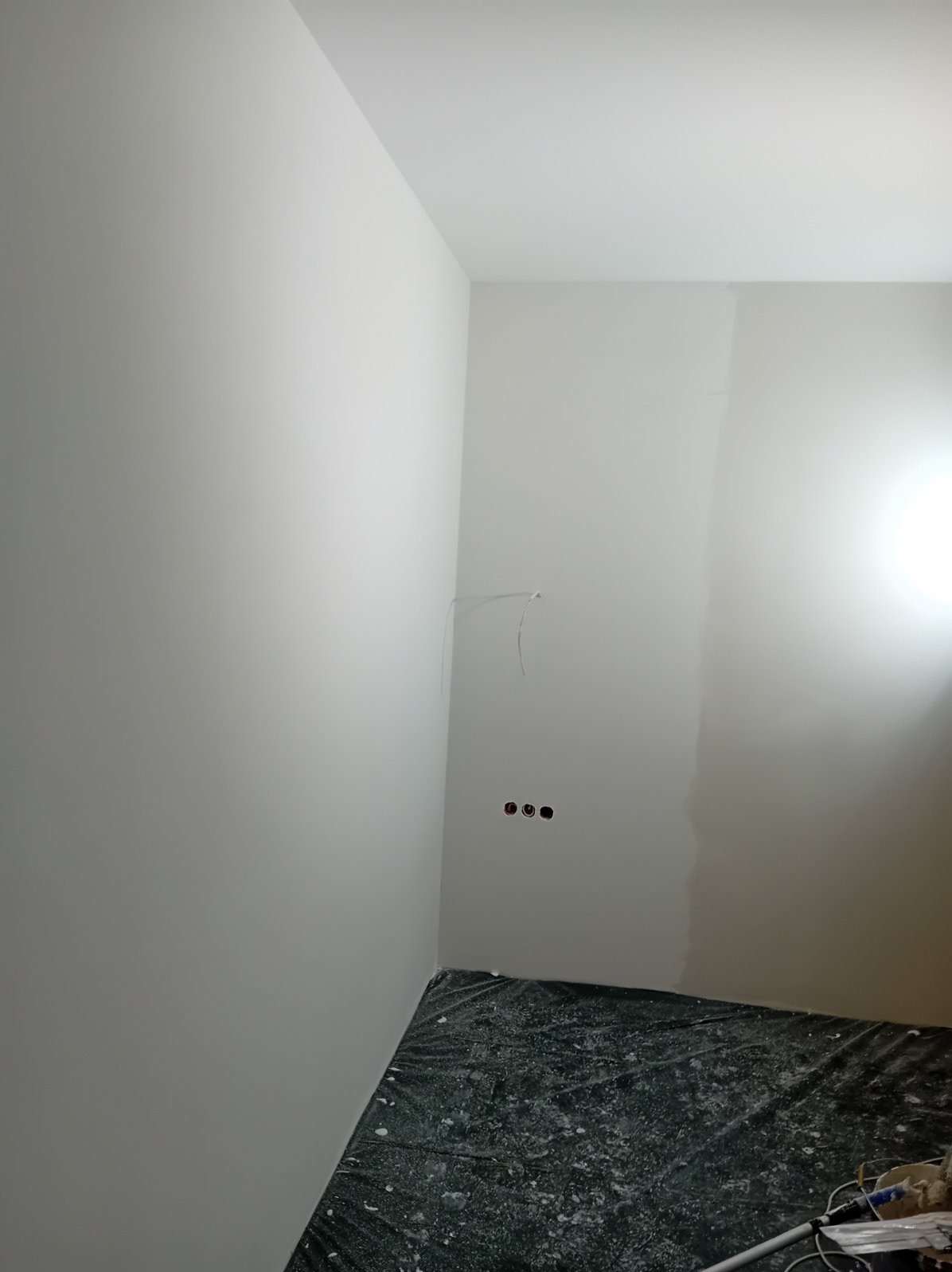 Świeżo pomalowane wnętrze pokoju na biało, z widocznymi trzema otworami na gniazdka elektryczne i wystającymi kablami, podłoga zabezpieczona folią malarską.
