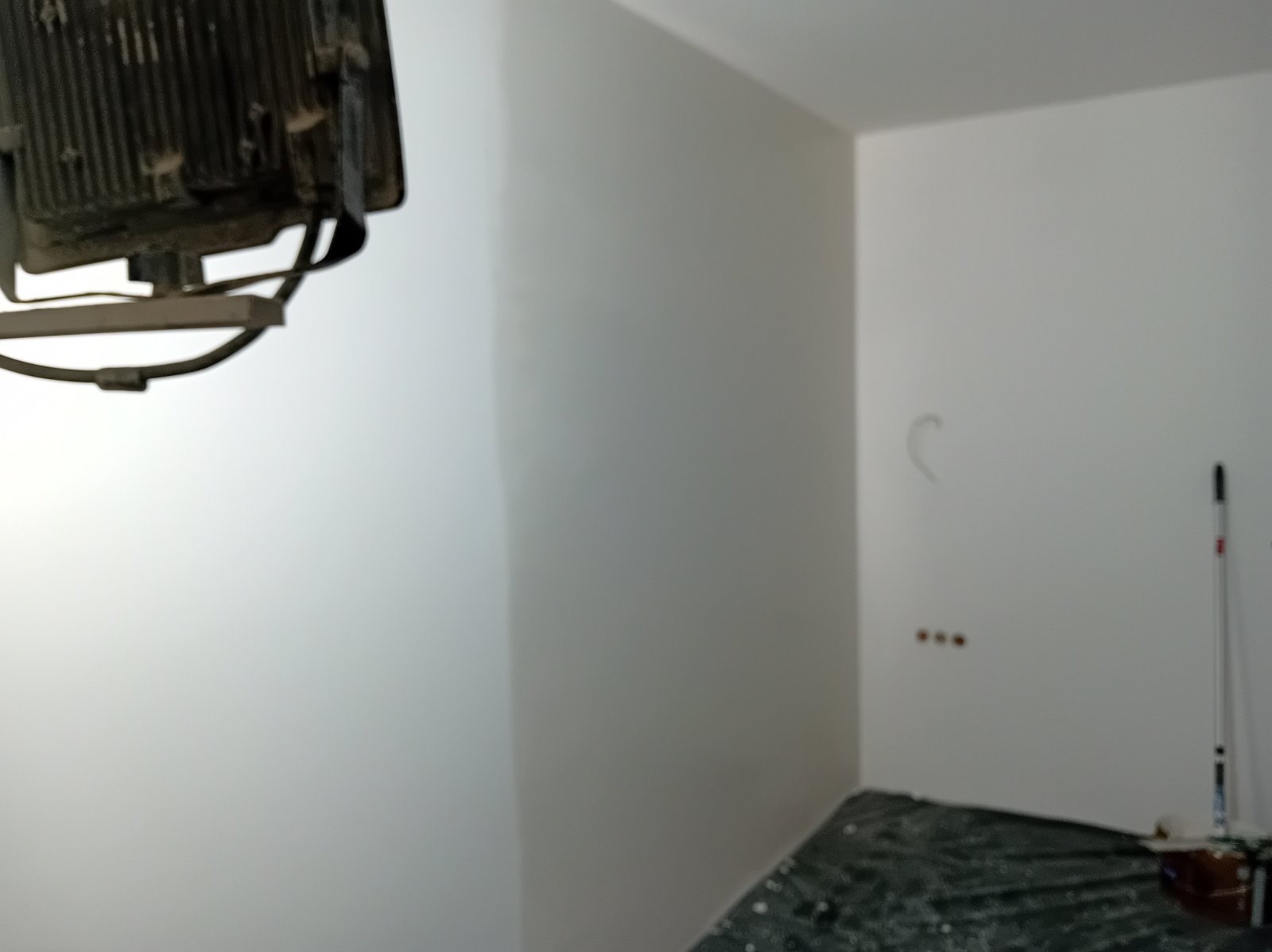 Przygotowane do malowania wnętrze z widocznym oświetleniem budowlanym, zabezpieczoną folią podłogą i śladami po gniazdkach na ścianie.