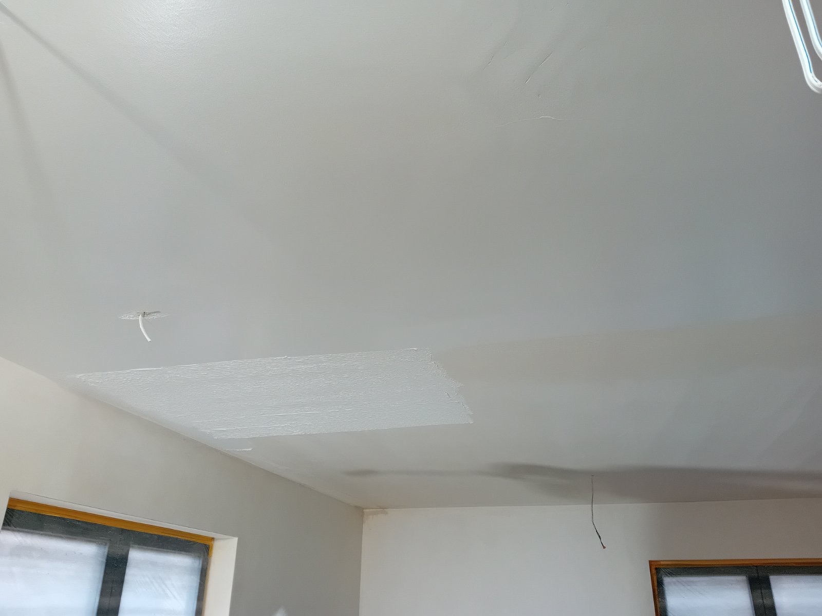Fragment sufitu po miejscowym nałożeniu gładzi, widoczna nierówna faktura świeżo nałożonego materiału, w tle okno zabezpieczone folią.