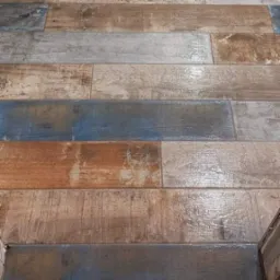 Podłoga wyłożona podłużnymi płytkami imitującymi drewno w odcieniach brązu, szarości i niebieskiego, ułożonymi poziomo, z widoczną fugą i fragmentem przypodłogowej listwy.