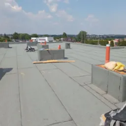 Widok z góry na płaski dach pokryty papą termozgrzewalną z detalami takimi jak kominki wentylacyjne, obróbki blacharskie i częściowo zużyte materiały budowlane, z widocznym krajobrazem miejskim...