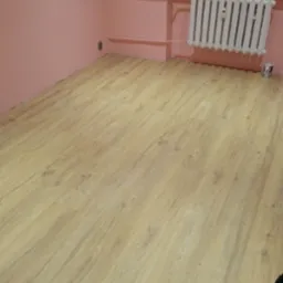 Ułożone panele podłogowe w pokoju z r&oacute;żowymi ścianami i białym grzejnikiem, widoczna puszka po farbie przy grzejniku.