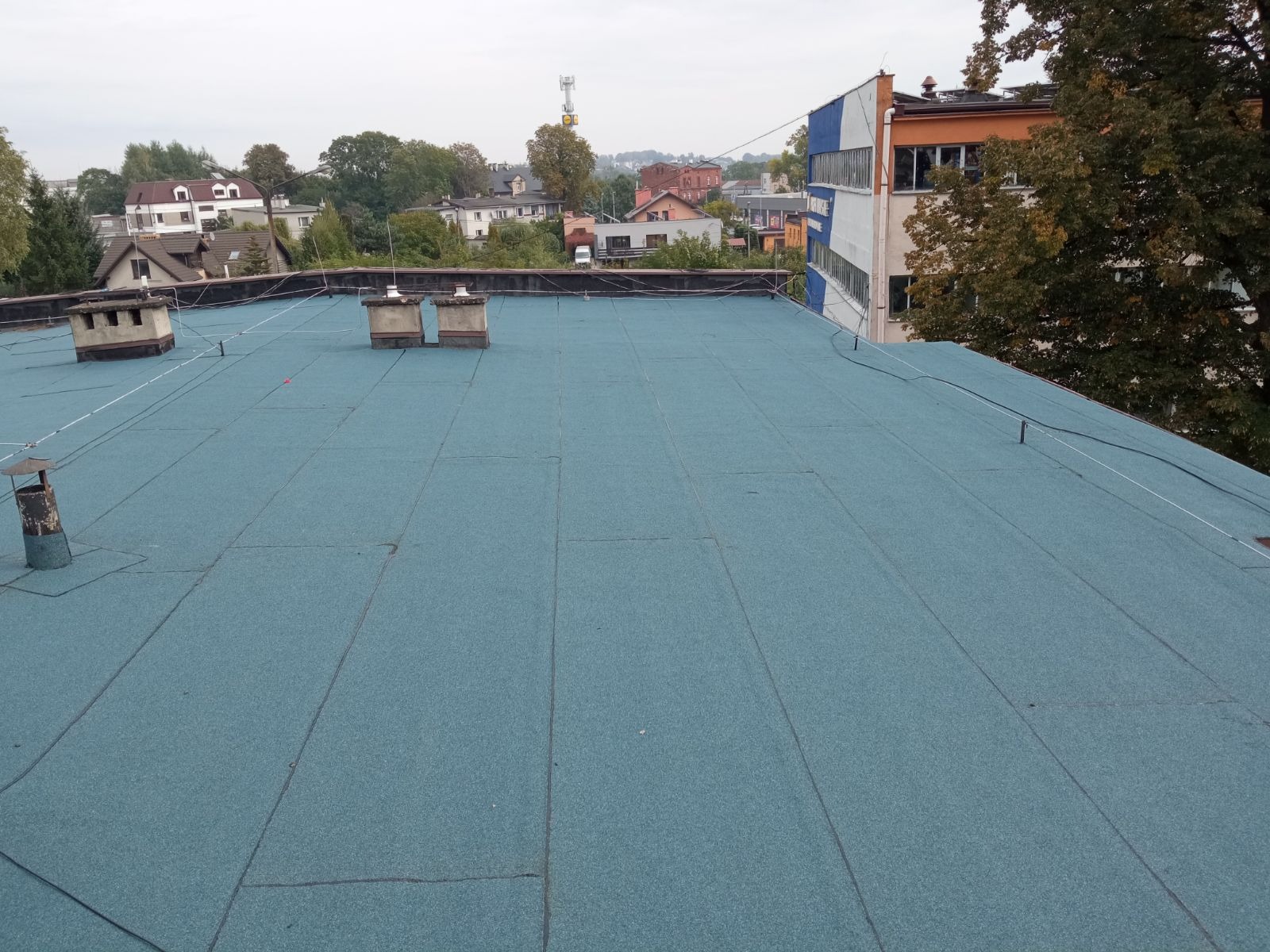 Błękitna, płaska powierzchnia dachu pokryta papą termozgrzewalną z widocznymi kominami wentylacyjnymi i przewodami odgromowymi na tle zabudowy miejskiej.